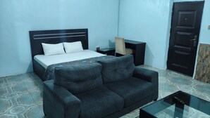 A bedroom with a bed, a sofa, a nightstand, a TV stand, and a door. - Oasis Place (Port Harcourt)