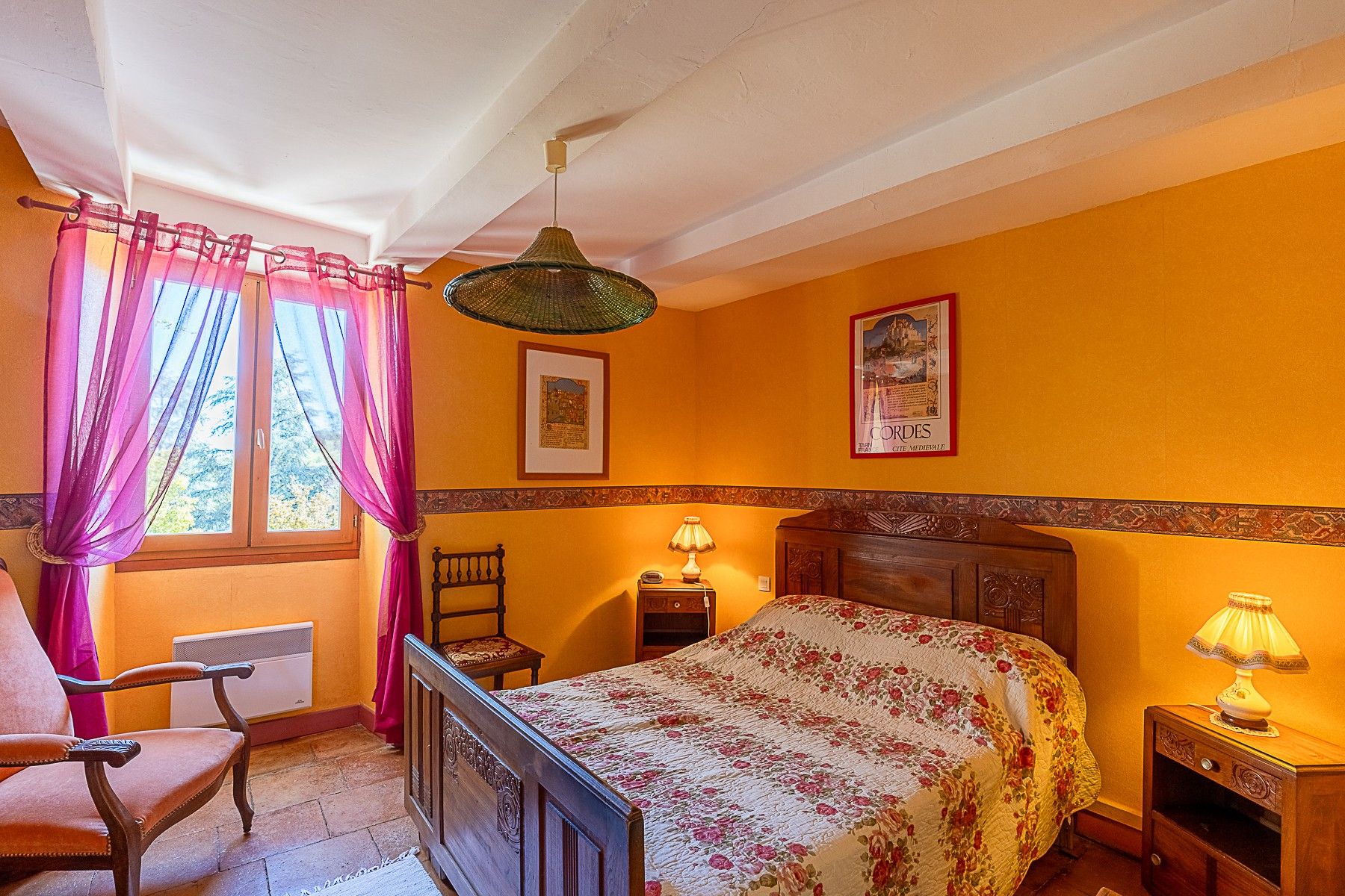 A bedroom with a bed, a chair, a nightstand, a lamp, a window with curtains, and a wall with a framed picture.