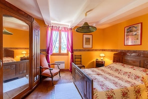 A bedroom with a wooden bed, a chair, a nightstand, a lamp, a window with curtains, and a framed picture on the wall.