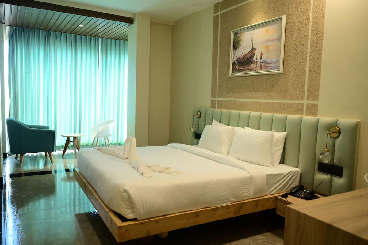 A hotel room with a large bed, a small desk, a chair, a painting on the wall, and a view of the outdoors through the window.