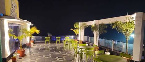 An outdoor terrace with green chairs and tables, potted plants, and a view of the city at night.