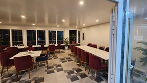 Ein Restaurant mit einem langen Tisch, roten Stühlen, einem gemusterten Boden und großen Fenstern.