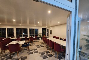 Ein Restaurant mit einem langen Tisch, roten Stühlen, einem gemusterten Boden und großen Fenstern.