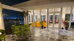 Eine Terrasse mit grünen Stühlen und einem Tisch, Kübelpflanzen und Blick auf die beleuchtete Stadt.