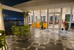 Eine Terrasse mit grünen Stühlen und einem Tisch, Kübelpflanzen und Blick auf die beleuchtete Stadt.