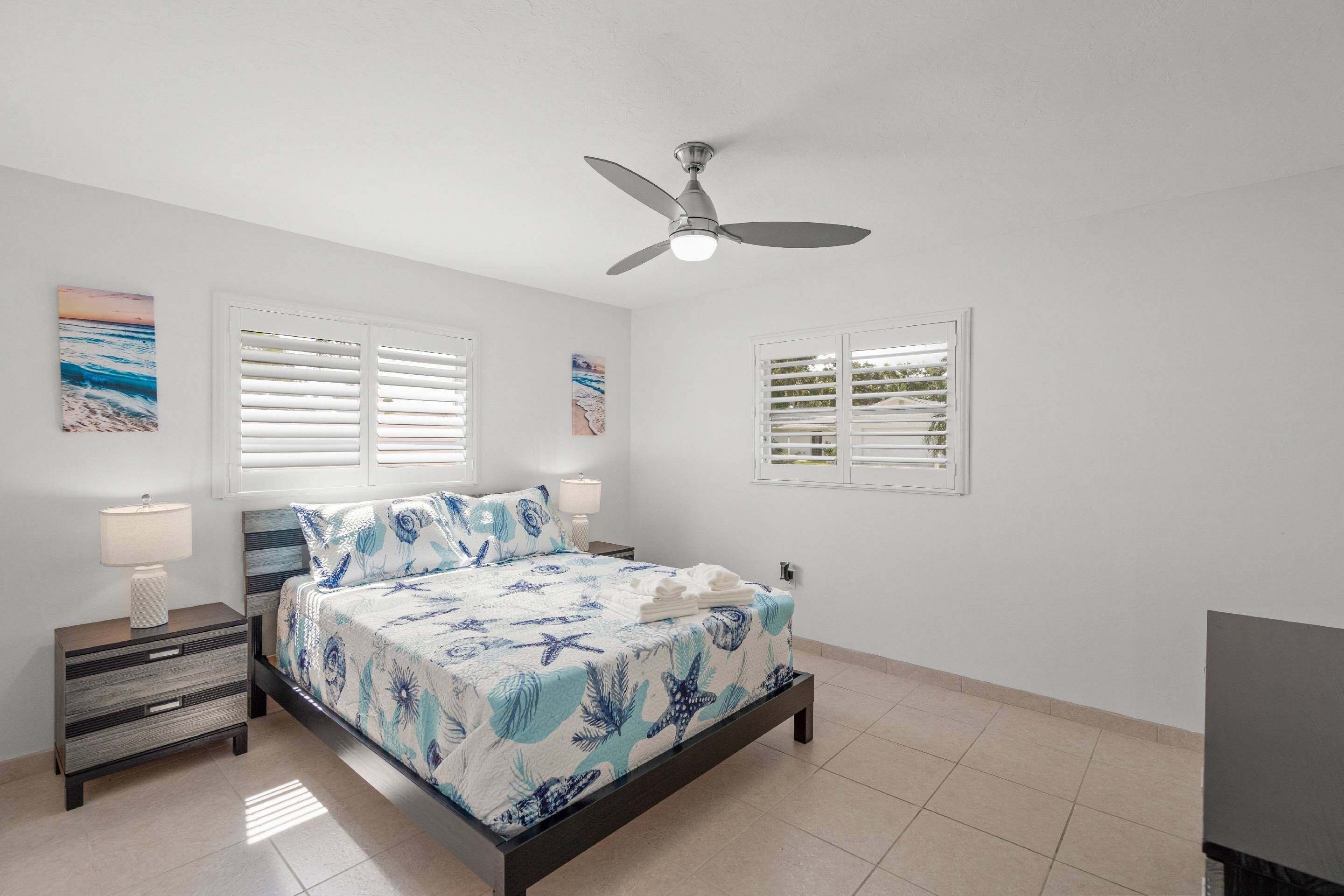 A bedroom with a bed, nightstands, a ceiling fan, and two windows with blinds.