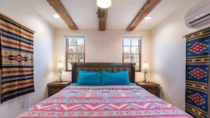 Ein Schlafzimmer mit einem Holzkopfende, zwei Nachttischlampen, einer gemusterten Bettdecke und einem Fenster mit Blick auf Bäume.