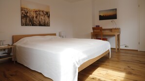 A bedroom with a large bed, a desk, and two chairs. There are paintings on the walls and a wooden floor.