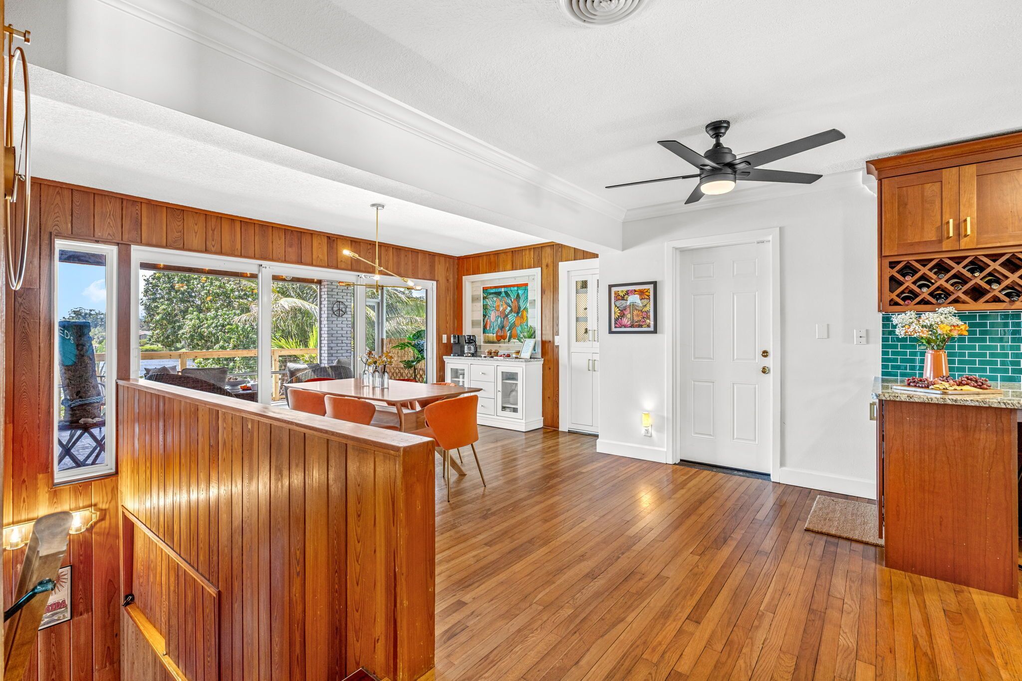 A modern kitchen with wooden cabinetry, a dining area with a wooden table and orange chairs, and a view of the outdoors through large windows.