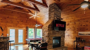 A spacious wooden cabin interior with a stone fireplace, a dining area, and a visible Netflix sign.