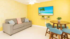 Ein Wohnzimmer mit einer Couch, einem Esstisch mit Stühlen, einem Deckenventilator und einem an der Wand hängenden Gemälde mit Strandmotiv.
