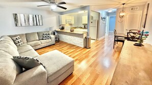 A modern living room with a sofa, wooden flooring, a ceiling fan, and a kitchen area with cabinets and appliances. - Recently Updated Trendy Biloxi Beach Condo (Biloxi)