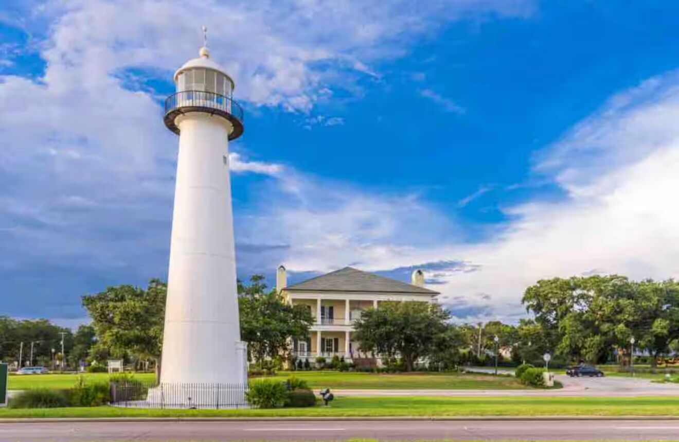 Un faro blanco con un balcón, una casa grande con porche y un cielo azul despejado.
