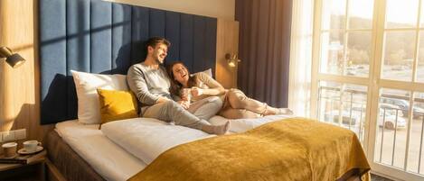Una habitación de hotel con una cama grande, una mesita de noche con una taza y un plato, y una ventana con vista a coches estacionados.