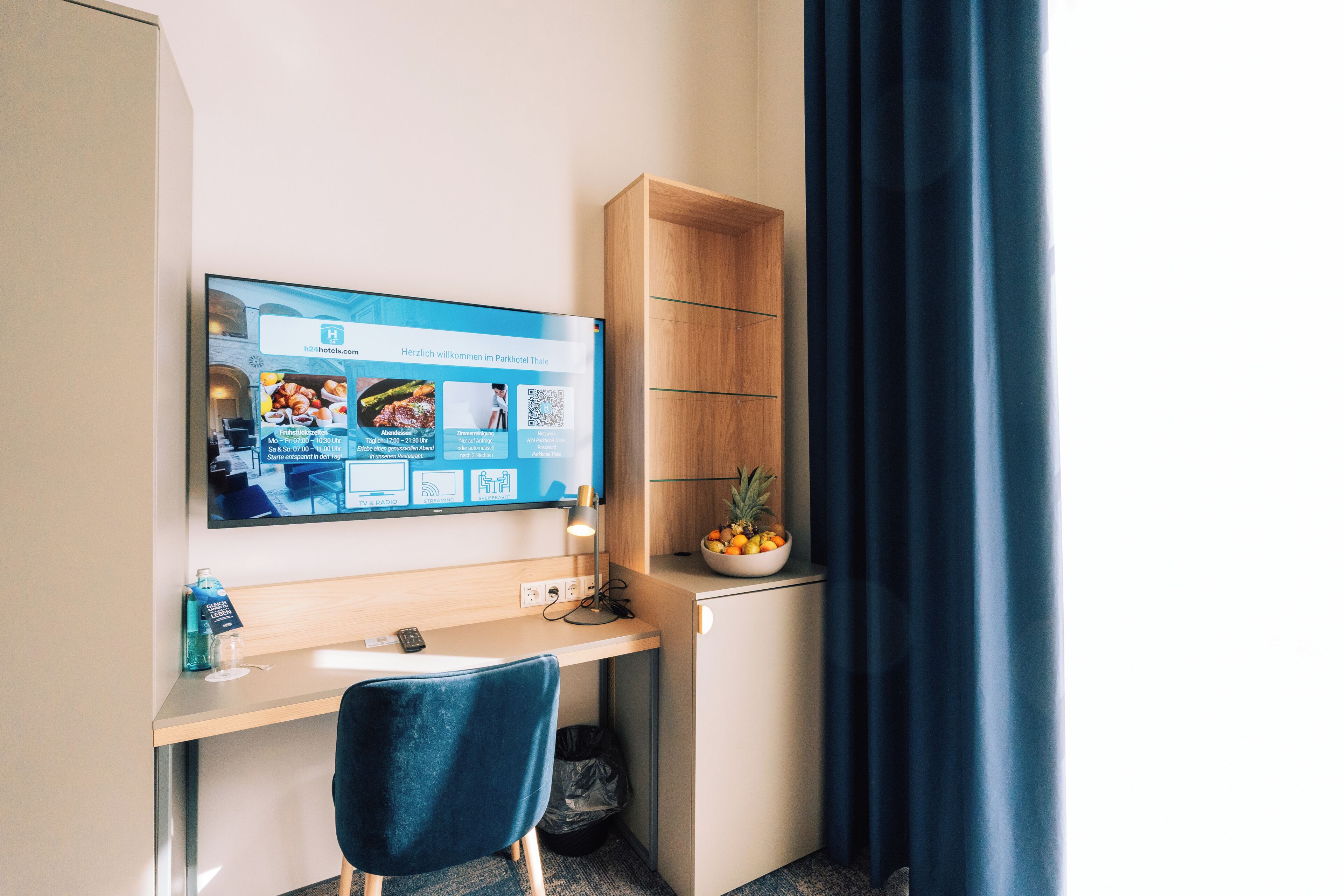 Habitación de hotel con escritorio, una silla, televisor mostrando la página web del hotel, un pequeño armario de madera con frutero y una cortina azul.