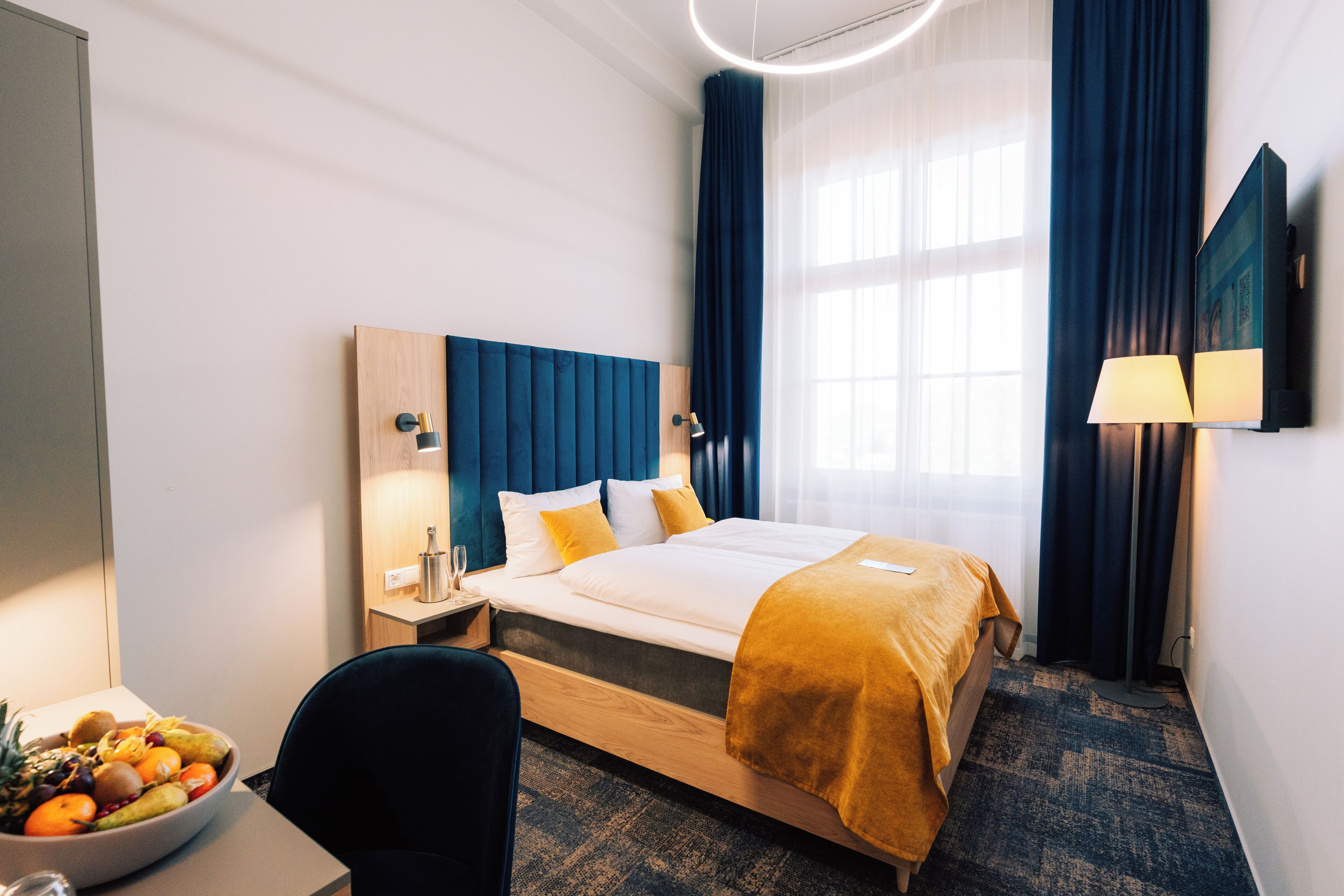 Una habitación de hotel con una cama, un escritorio con un frutero, una silla, una mesita de noche y una lámpara.