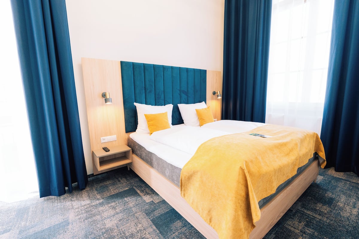 A hotel room with a bed, a nightstand, a lamp, and a window with curtains.