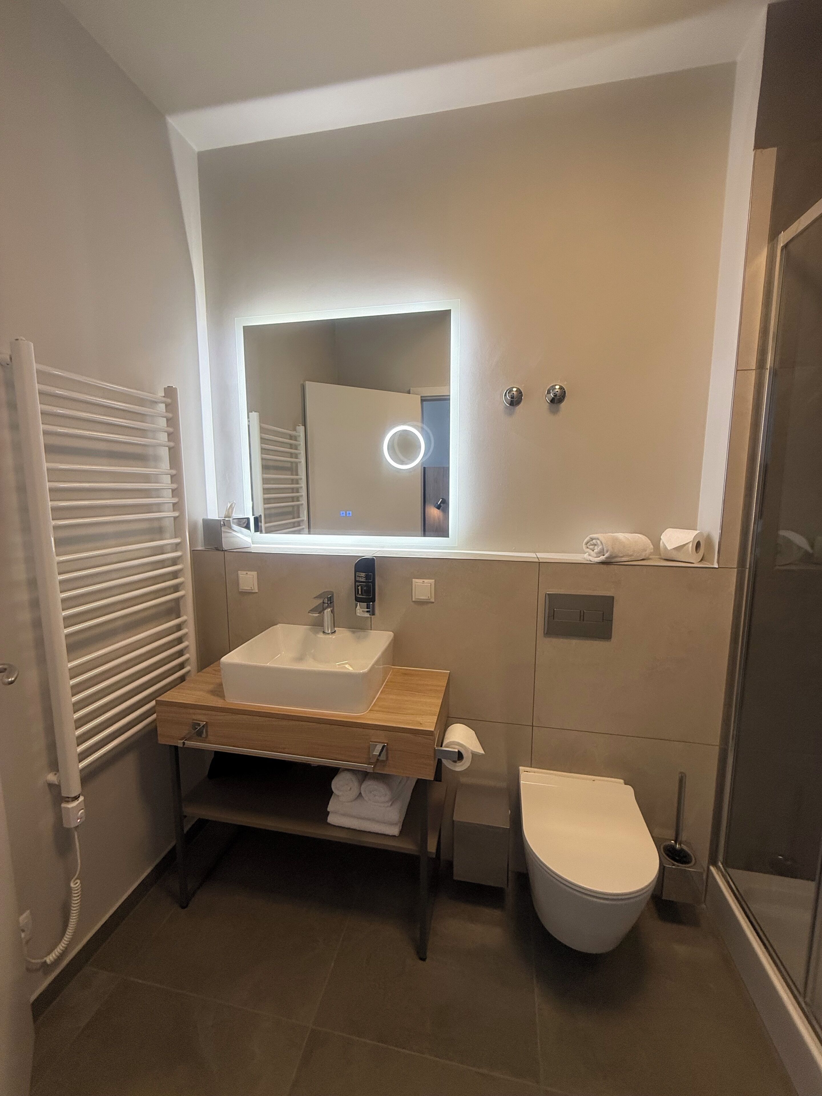 A modern bathroom with a wooden vanity, a white sink, a mirror with a ring light, a toilet, and a shower area.