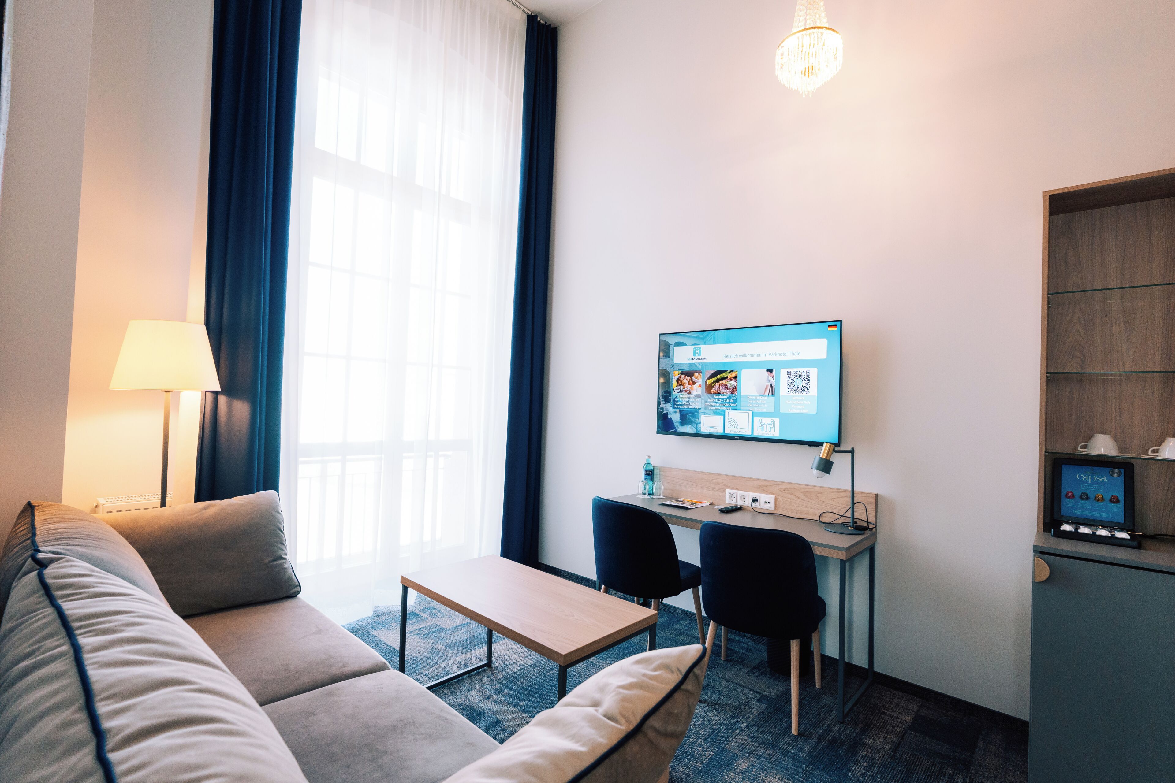 A hotel room with a sofa, a small table, a desk with a computer, and a mini-fridge.