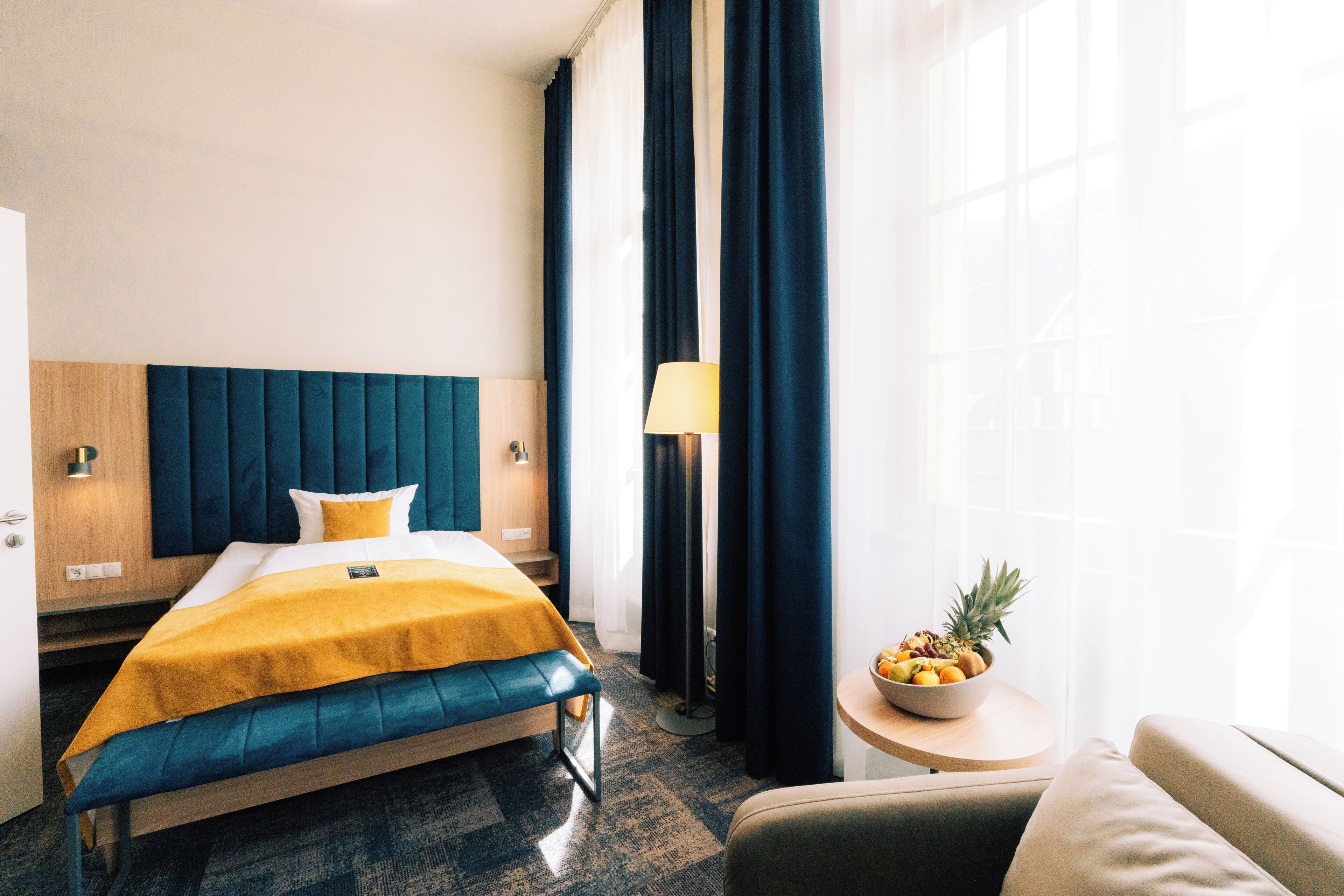 A hotel room with a bed, a bedside table with a lamp, a small round table with a fruit bowl, and a window with curtains.
