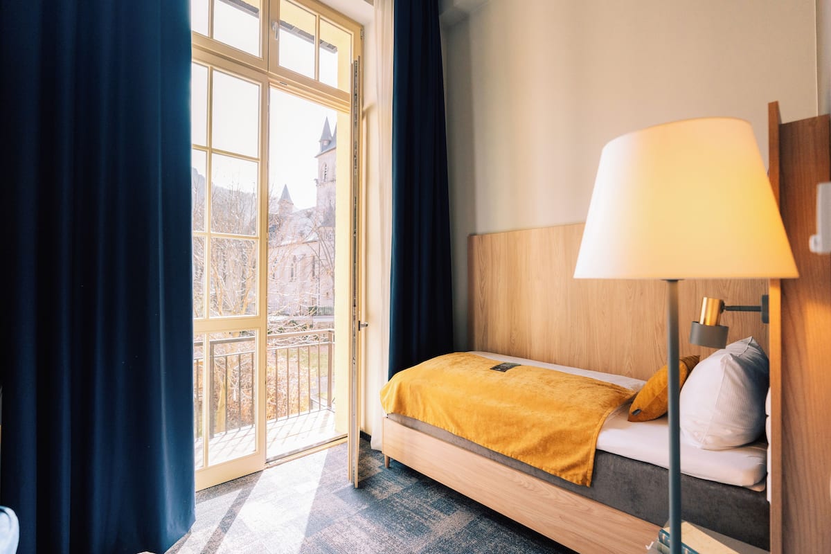 A hotel room with a bed, a lamp, a window with curtains, and a view of a building outside.