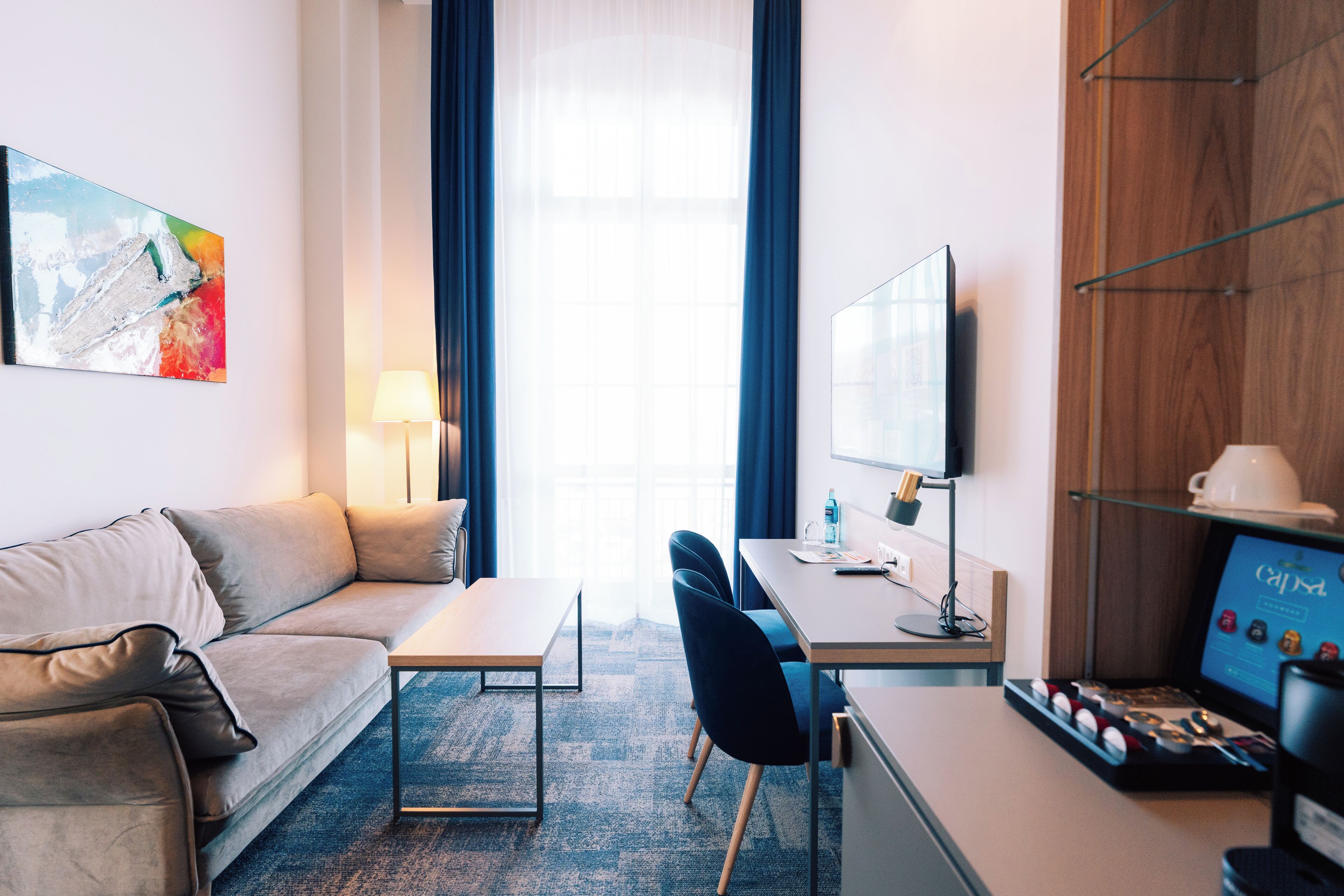 Una habitación de hotel moderna con sofá, escritorio, silla, máquina de café y un amplio ventanal con cortinas.