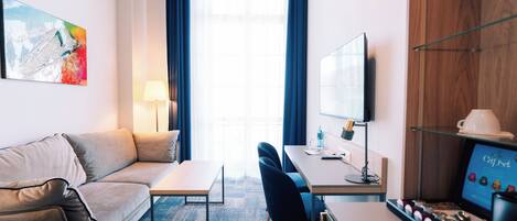 Una habitación de hotel moderna con sofá, escritorio, silla, máquina de café y un amplio ventanal con cortinas.