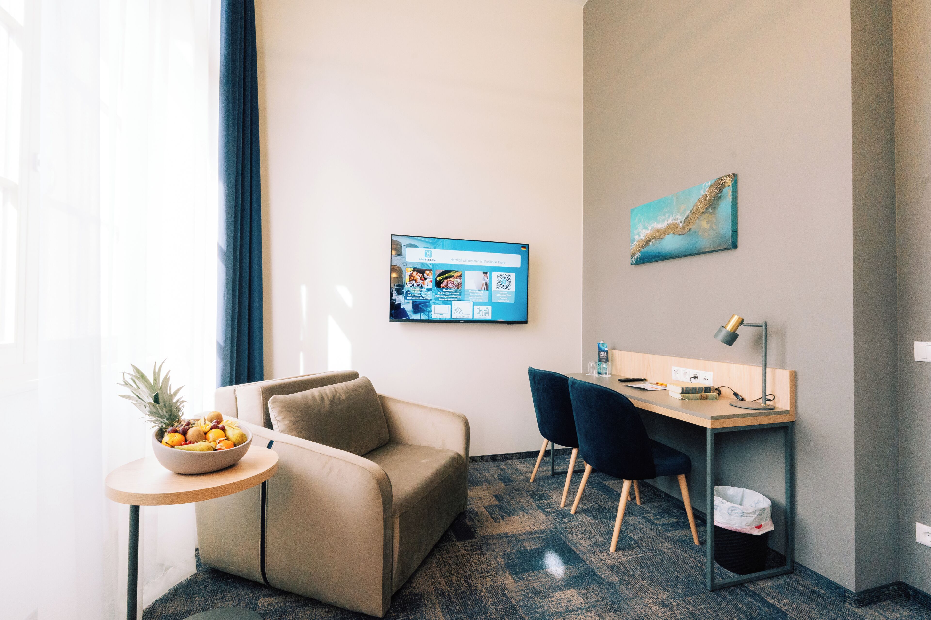 A hotel room with a sofa, a desk with a computer, a small table with fruit, and a wall-mounted TV.