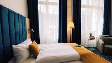 Una habitación de hotel con cama, mesita de noche, lámpara, silla y ventana con cortinas.
