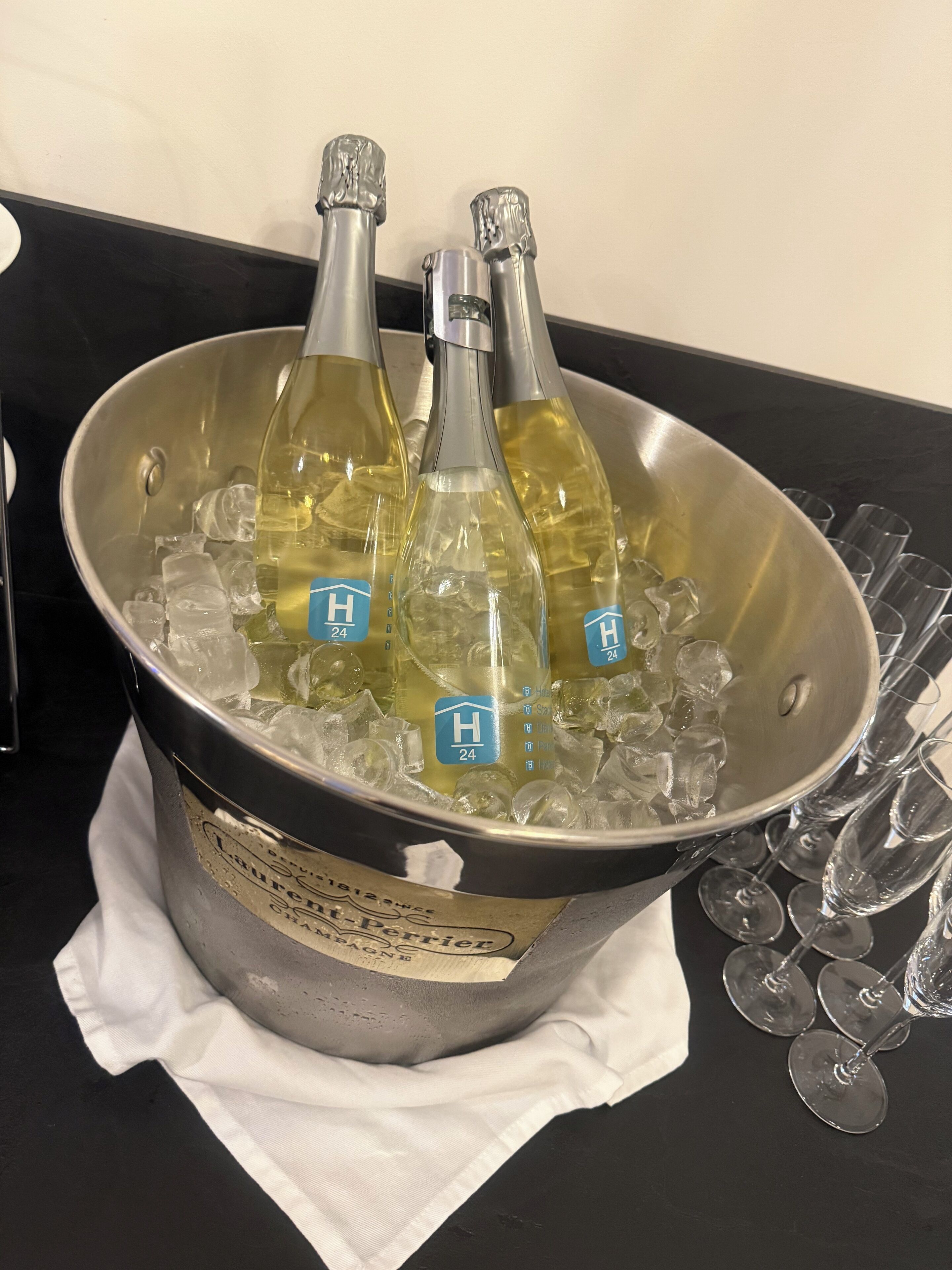 A bucket filled with ice and three bottles of wine, placed on a table with several empty wine glasses around it.