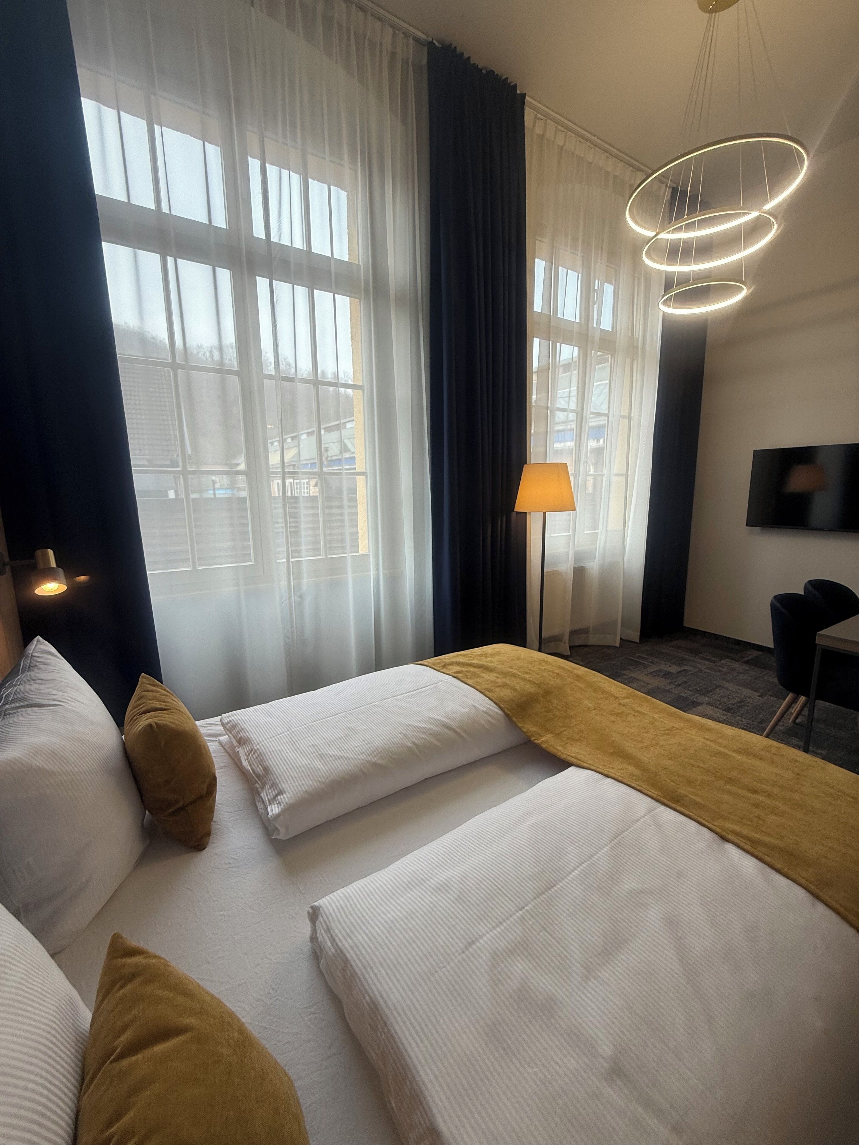 A hotel room with two beds, a desk, a TV, and a large window with curtains.