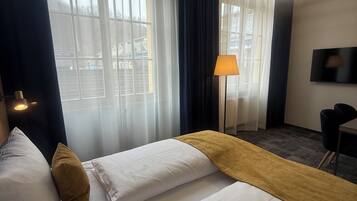 A hotel room with two beds, a desk, a TV, and a large window with curtains.