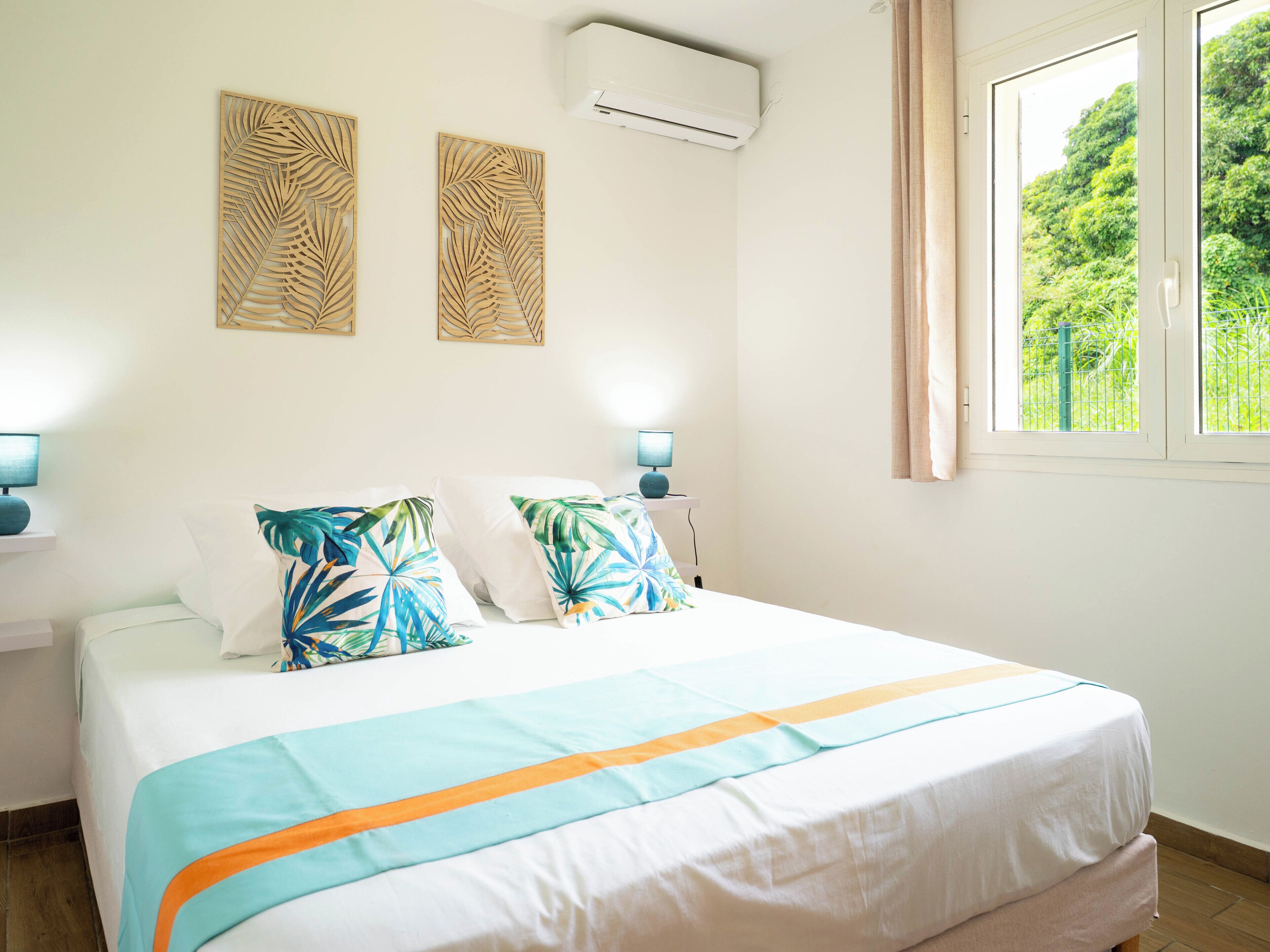 A bedroom with a bed, two wall-mounted air conditioning units, a window with a view of greenery, and two wall-mounted lamps.