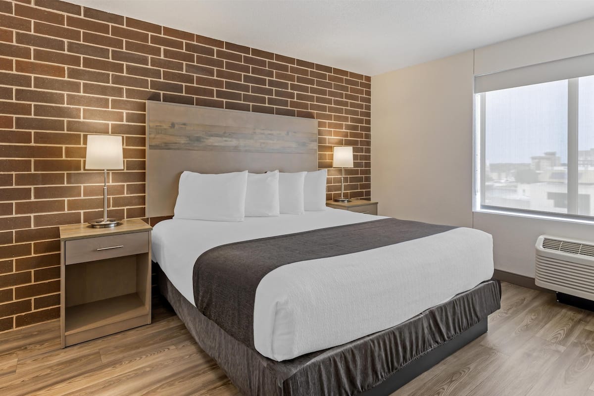 Una habitación de hotel con pared de ladrillo, una cama grande con ropa blanca, dos lámparas de noche y una ventana con vistas a la ciudad.