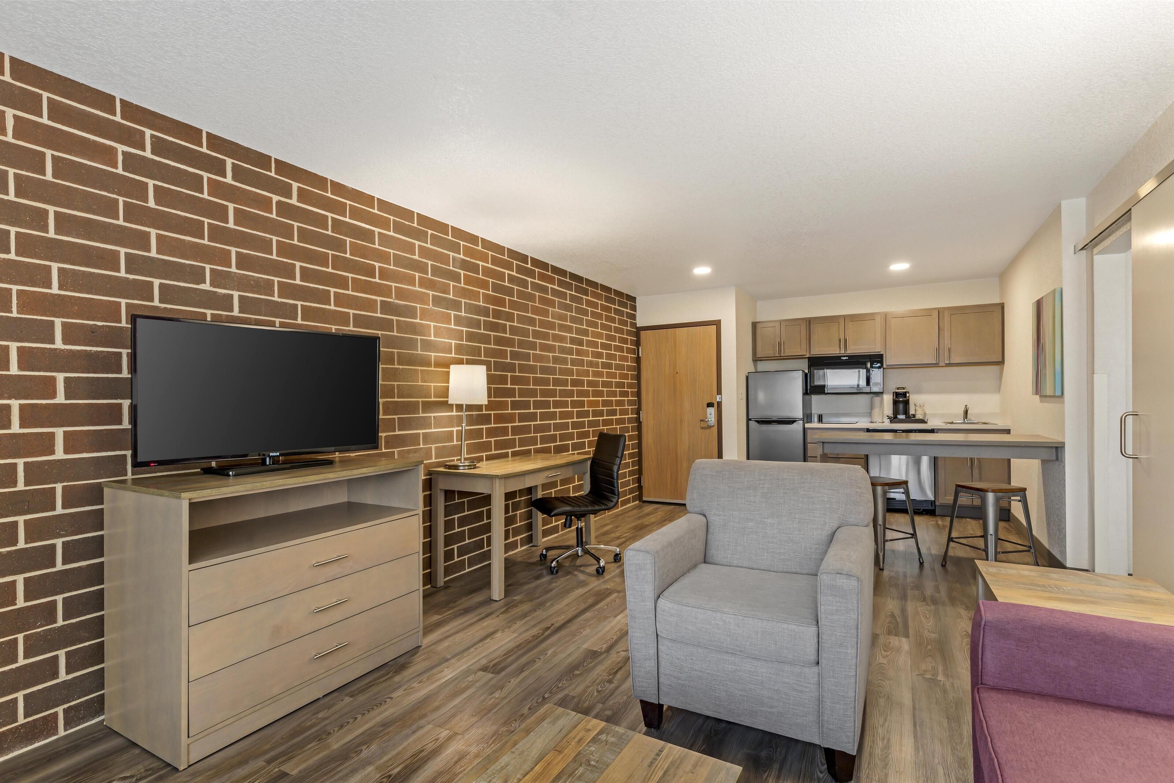 A modern living room with a flat-screen TV, a sofa, a desk with a chair, and a kitchen area with cabinets and appliances.