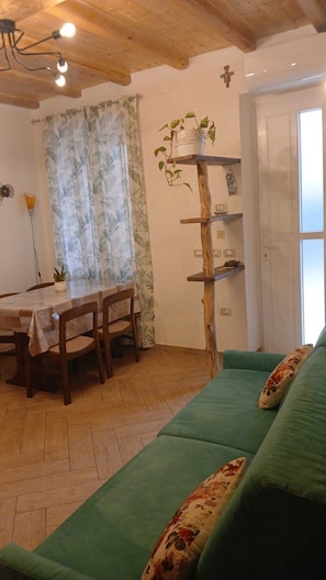 Ein gemütliches Zimmer mit Holzbalkendecke, einem Esstisch mit Stühlen, einem grünen Sofa und einem Holzregal mit einer Pflanze.