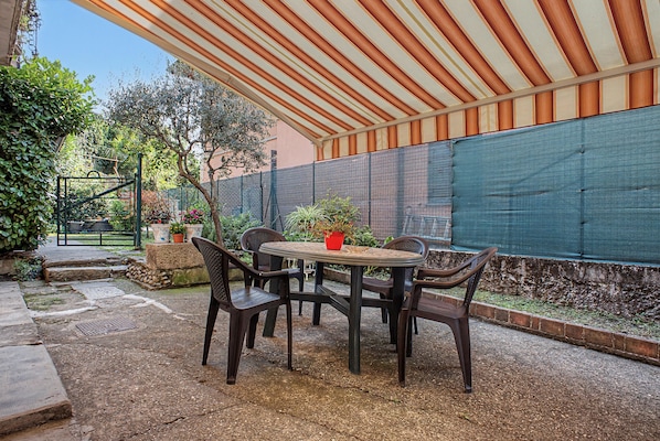 Un patio exterior con una mesa y sillas, rodeado por una zona cubierta y una cerca verde.
