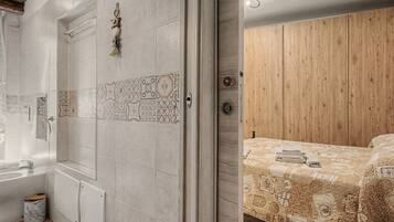 Un bagno con doccia, WC e lavandino. Una porta in legno conduce a una camera da letto con un letto e un'armadio in legno.