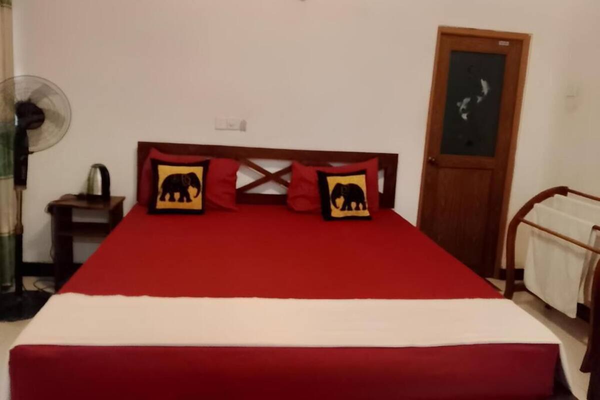 A bed with a red bedspread and two pillows with elephant designs, a wooden headboard, a wooden door, and a small table with a kettle.