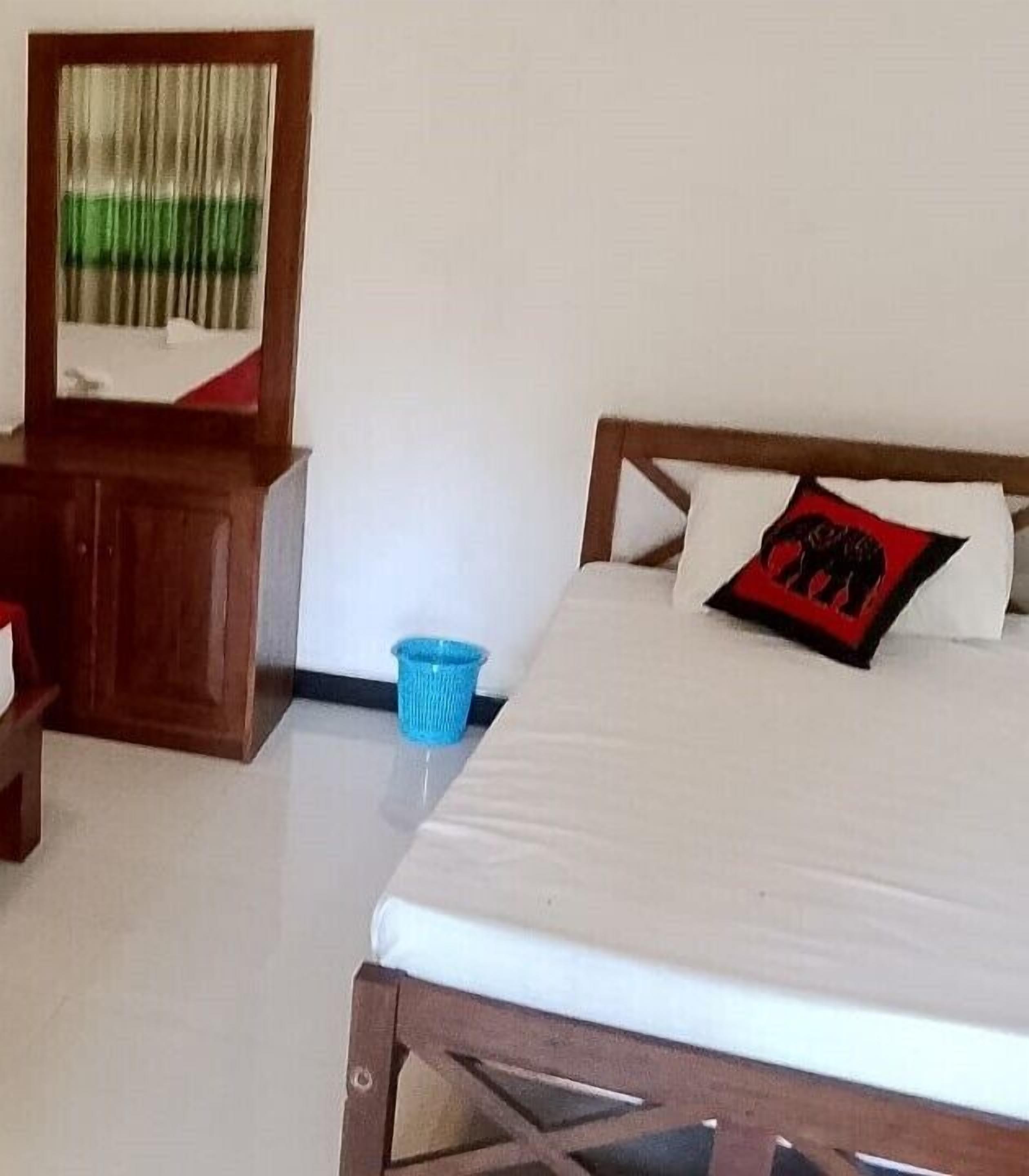 A bedroom with a wooden bed frame, a white mattress, a blue trash bin, a wooden nightstand, and a mirror with a green and white curtain.