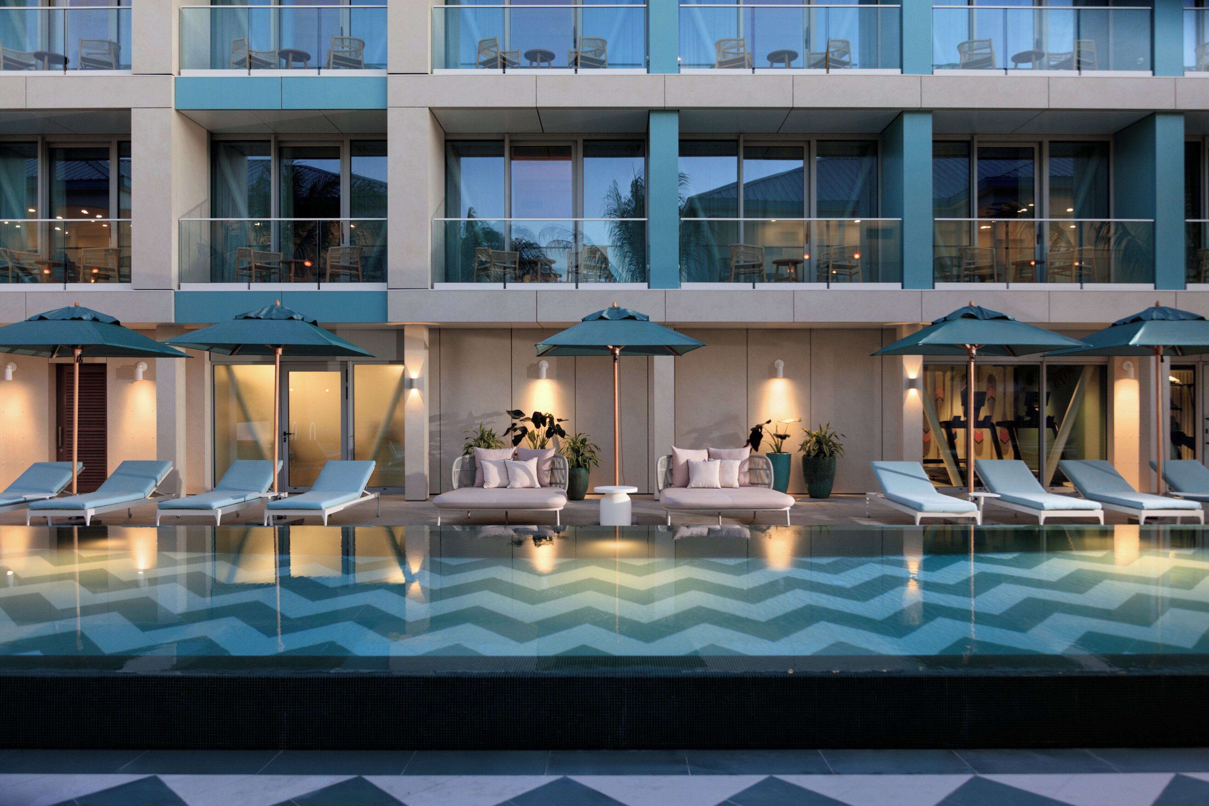Un hotel moderno con piscina, sillones de descanso y mobiliario exterior con sombrillas.
