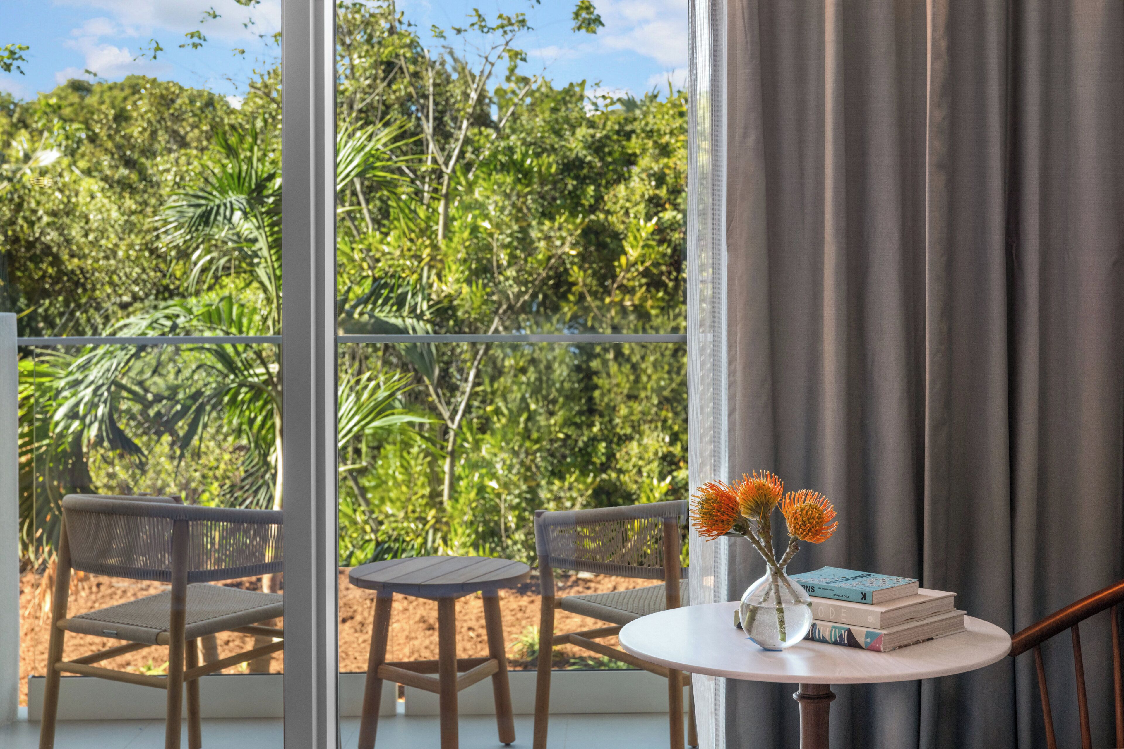 A balcony with two wicker chairs, a small round table with a vase of flowers and books, and a view of dense greenery.