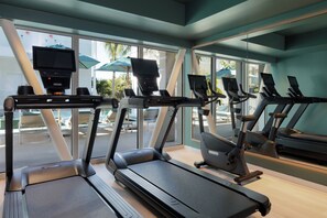 Ein Fitnessraum mit Laufbändern, Crosstrainern und einem großen Fenster mit Blick auf einen Poolbereich.