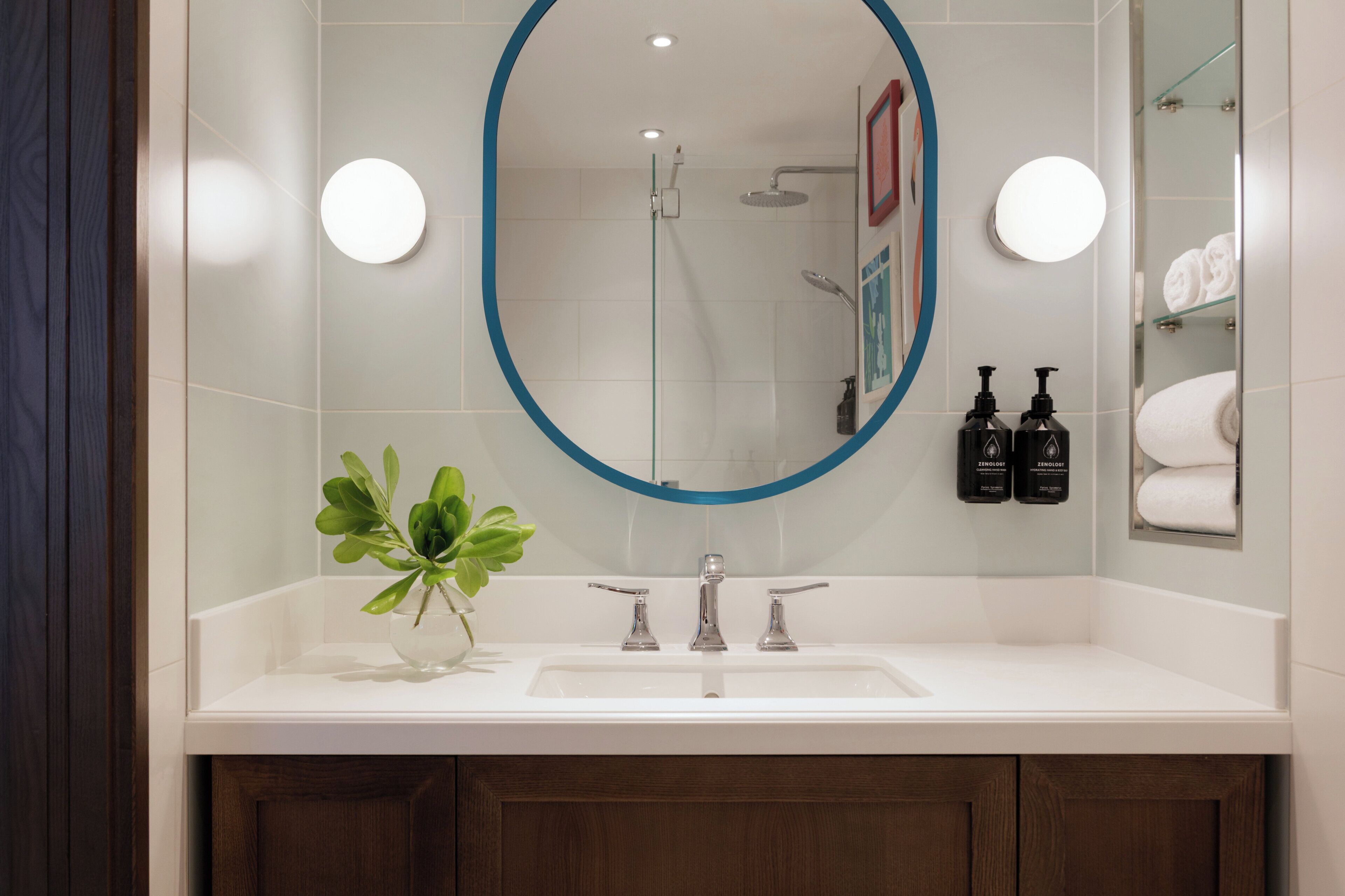 A bathroom with a white sink, a mirror with a blue frame, a shower, and a glass shelf with towels.