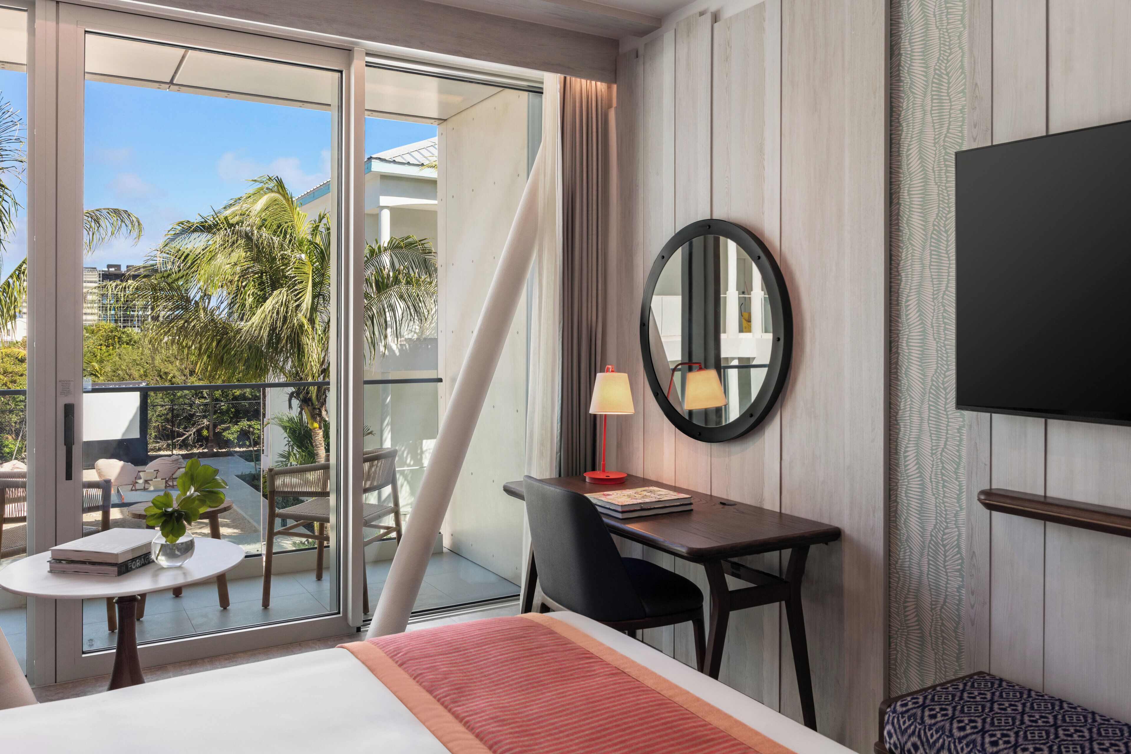 Quarto de hotel com cama, escrivaninha com luminária, cadeira, pequena mesa com planta e televisão na parede.