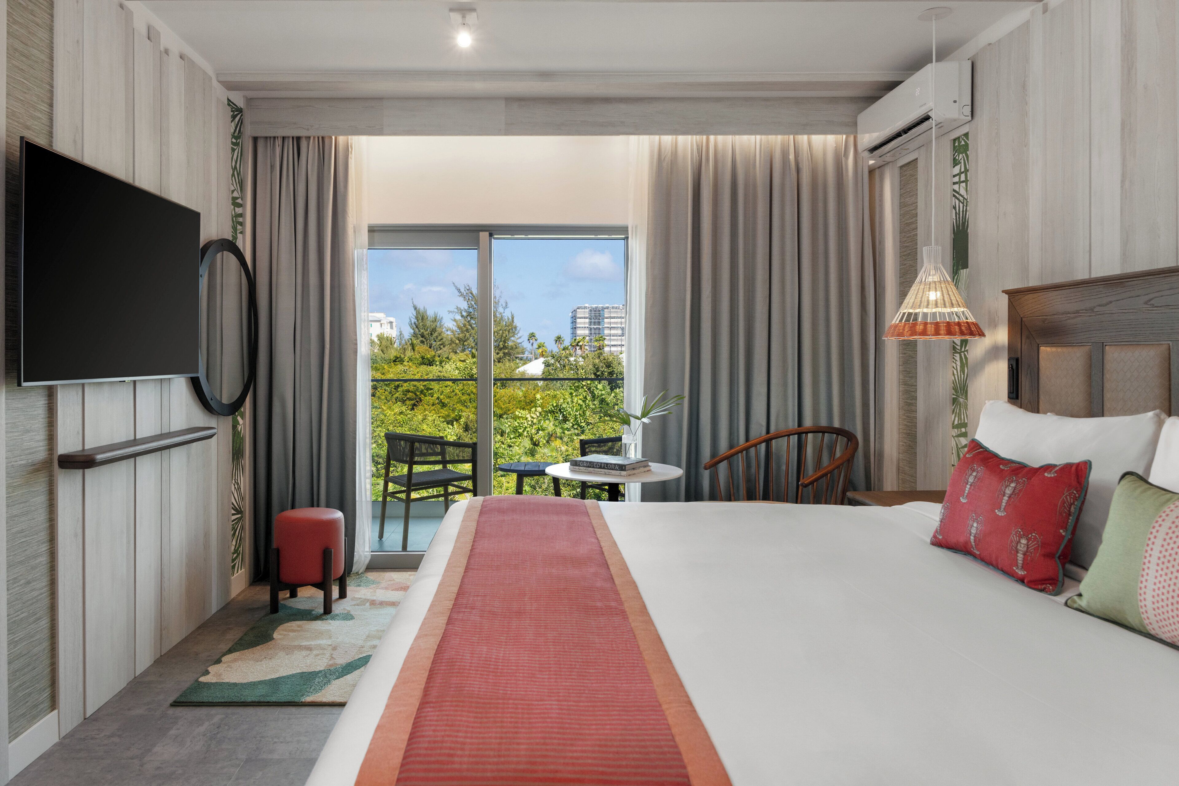 Habitación de hotel con cama, televisor, balcón con vista a edificios y follaje, una pequeña mesa con una silla y una lámpara colgante.