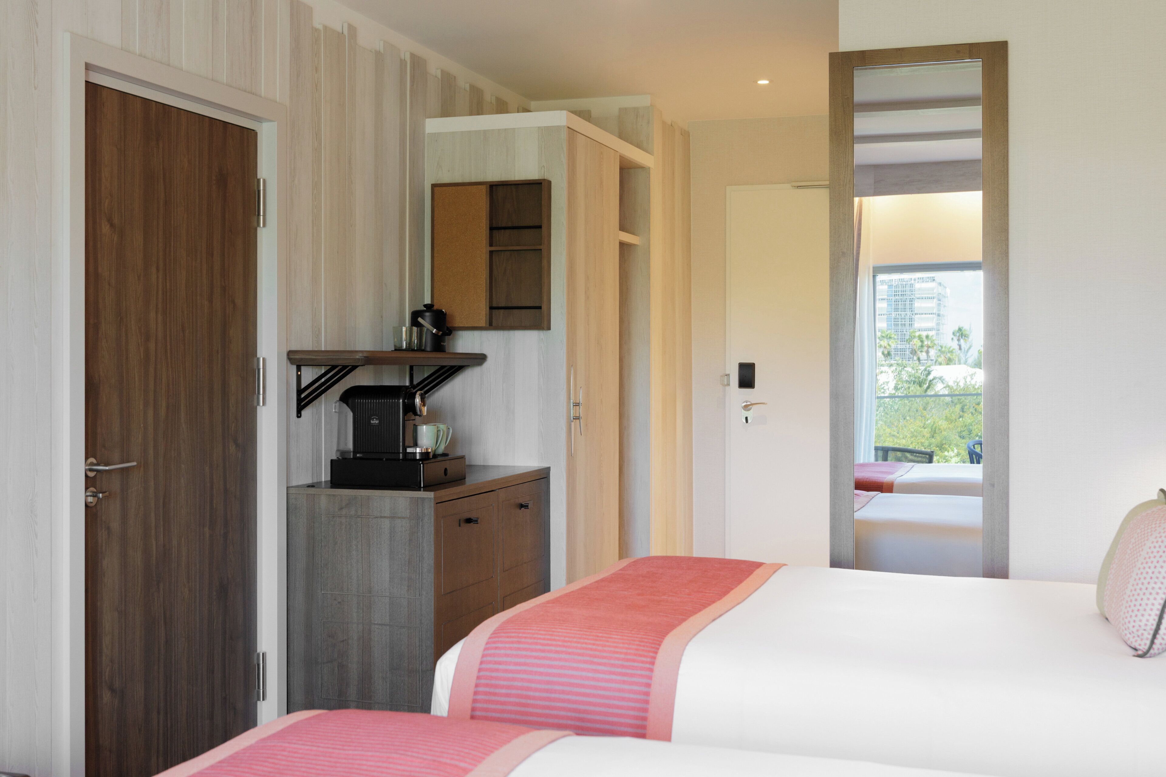 A hotel room with a bed, a desk with a coffee machine, a mirror, and a view of the outdoors.