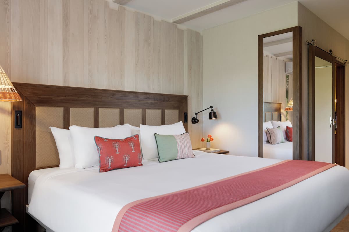A hotel room with a large bed, wooden headboard, bedside tables, a nightstand, a mirror, and a wall-mounted lamp.
