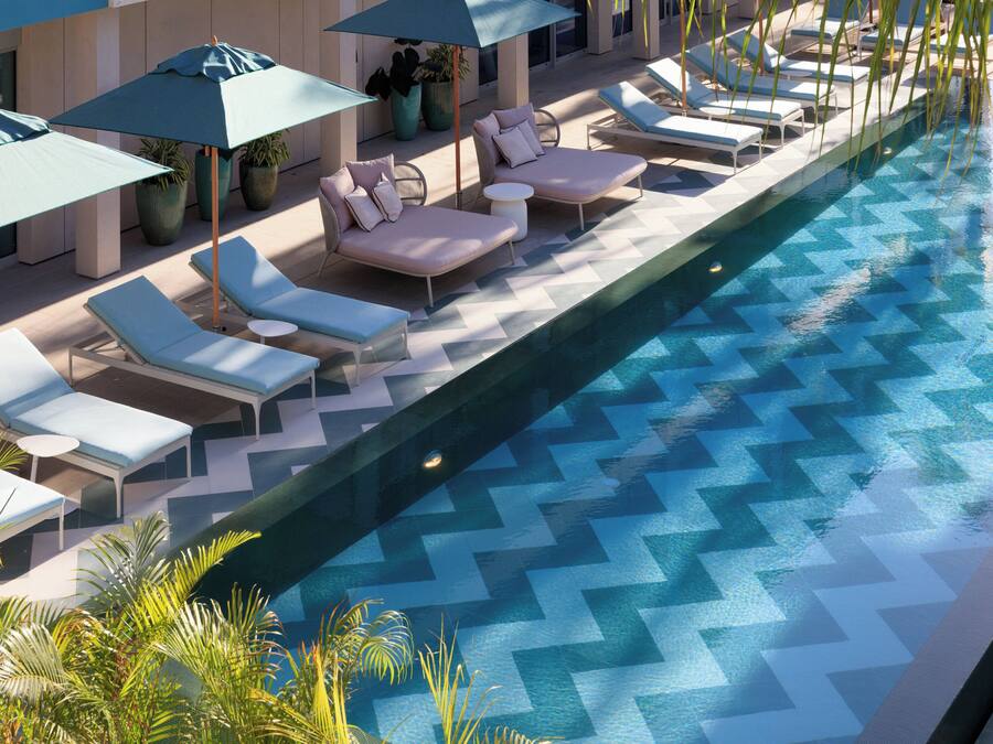 Zona de piscina con sillas de descanso y sombrillas, rodeada de plantas en macetas.