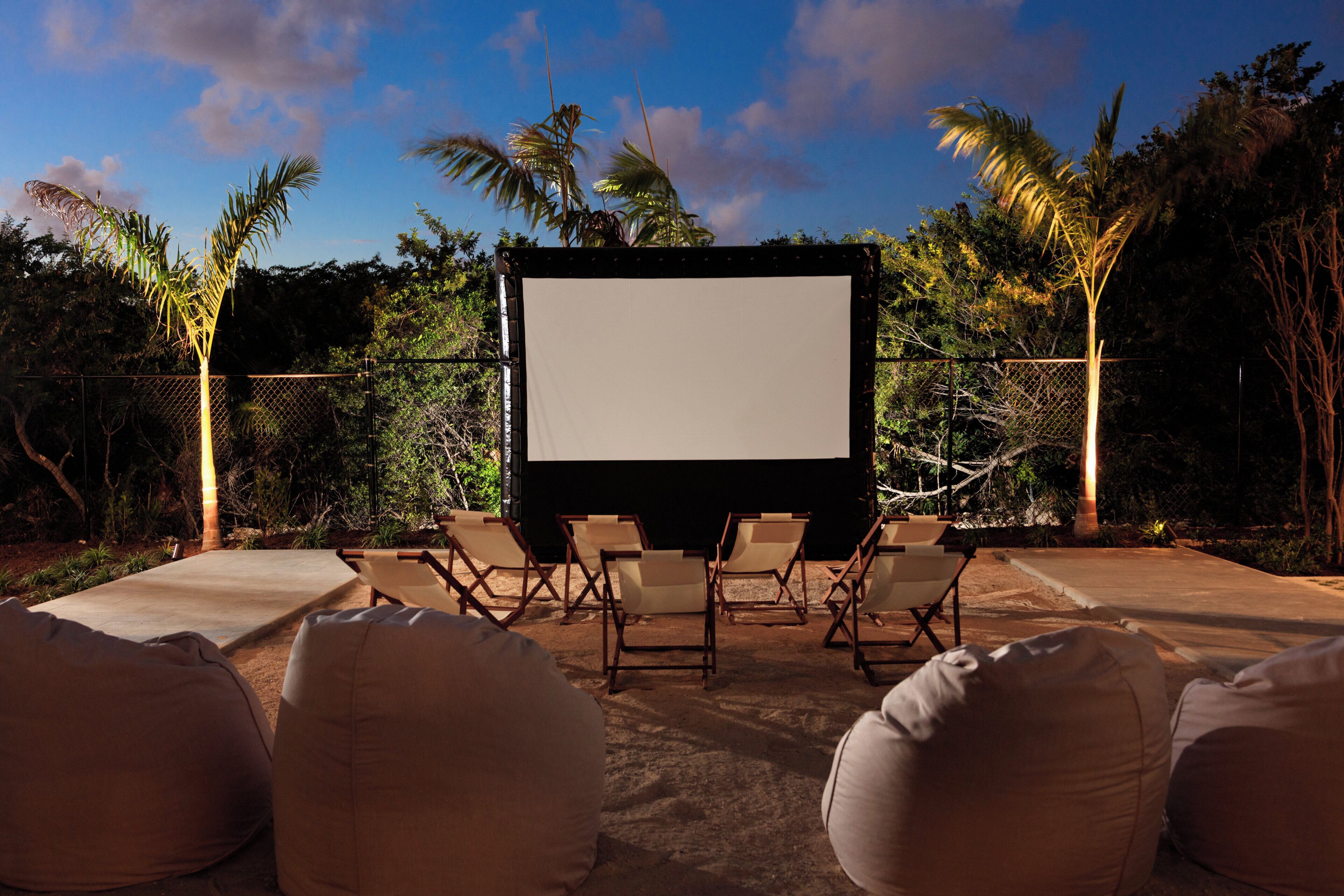 Un écran de cinéma en plein air, sur lequel rien n’est projeté pour le moment, entouré de fauteuils et de poufs, avec en arrière-plan des palmiers et un ciel dégagé.